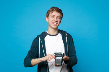 Joyful young man holding wireless modern bank payment terminal to process and acquire credit card payments isolated on blue background. People sincere emotions, lifestyle concept. Mock up copy space.