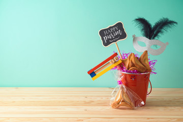 Jewish holiday Purim background with bucket, carnival mask, noisemaker and hamantaschen cookies on wooden table.
