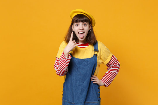 Funny girl teenager in french beret showing tongue, horns up gesture, depicting heavy metal rock sign, rock-n-roll isolated on yellow background. People emotions lifestyle concept. Mock up copy space.
