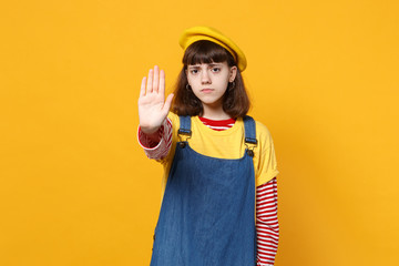 Portrait of upset girl teenager in french beret, denim sundress showing stop gesture with palm isolated on yellow background in studio. People sincere emotions, lifestyle concept. Mock up copy space.