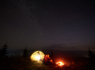 Romantic couple tourists boy and girl sitting near illuminated tent by burning bonfire under starry sky, enjoying quiet night camping in mountains. Tourism, traveling and beauty of nature concept.
