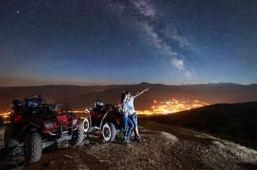 Young family man and woman tourists with atv quad motorbikes on the top of mountain. Man pointing at beautiful night sky full of stars, Milky way, luminous town on background © anatoliy_gleb
