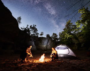 Young couple travellers man and woman resting beside camp, campfire and glowing tourist tent at night. On background beautiful night starry sky full of stars and Milky way, mountain rocks, trees