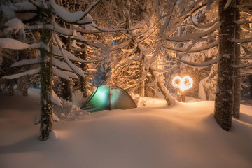 Night shot of burning fire, long exposure, sleeping in the snow outside. Night bivouac in the mountains, millions of hours under the night sky, green lighted tent in the Alps.