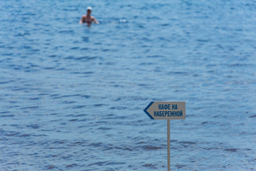 The inscription on the sign-plate "Cafe on the waterfront." A woman swims in the sea or river. Summer Sunny day on the beach.
