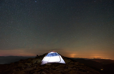 Naklejka na ściany i meble Tourist camping at summer night on the top of rocky mountain. Glowing tent under amazing night sky full of stars. On background starry sky, mountains and luminous town. Tourism adventure concept