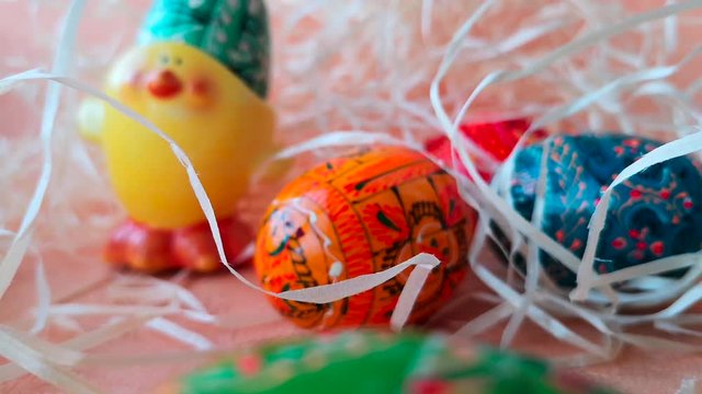 Happy Easter concept: multicolored painted Easter eggs and ceramic egg holder made in the shape of a chicken among decorative straw. The straw moves slightly in the wind.