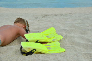 A cheerful and funny boy in flippers and a mask for scuba diving lies on the sand by the sea and sunbathes in the sun in a clear summer.
