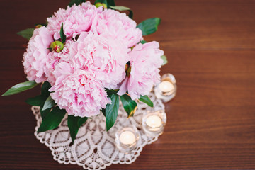 A bouquet of beautiful pink peony flowers in a vase on a table surrounded by candles.  Copy space