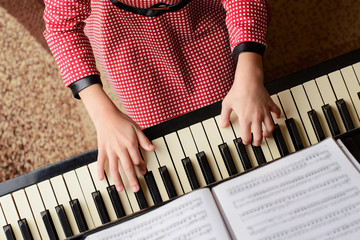 Little girl in red dress performing classical music at home playing the piano looking to the music...