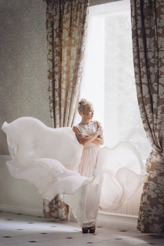 Cute blonde girl in the image of the bride stands near the window in a flying bright dress. wedding photo. gentle calm image. Small Noise