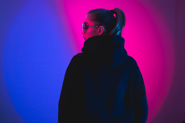 Portrait of fashion young girl in a black sweater with a hood and round sunglasses in red and blue neon light in the studio - obrazy, fototapety, plakaty