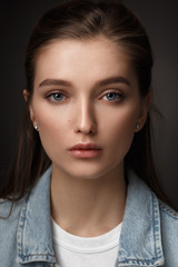 Portrait of beautiful brunette girl with hair tied back dressed in jeans jacket on the dark background in the studio