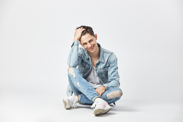 Smiling dark-haired guy dressed in a white t-shirt, jeans and a denim jacket is sitting cross-legged on the floor on the white background in the studio