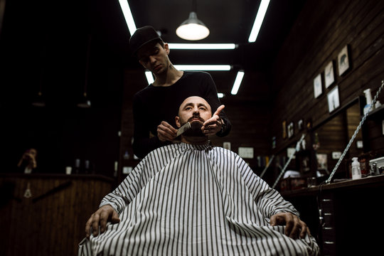 The stylish barbershop. The fashion barber tidies up beard of brutal man sitting in the armchair