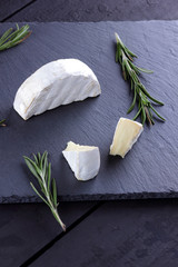 Camembert and rosemary on black stone board. Soft cheese with white mold on black background. Sliced cheese and rosemary branches on slate board