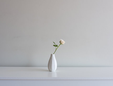 Close up of single pale pink rose in small vase in the centre of white sideboard against neutral wall background