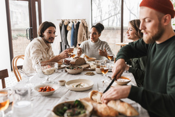 Handsome man in hat with beard sitting at the table thoughtfully cutting baguettes. Group of young international friends having lunch spending time together in cozy cafe