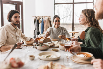 Pretty african american woman with dark curly hair sitting at the table with soup and slice of bread in hand happily looking in camera. Group of young international friends having lunch in cozy cafe