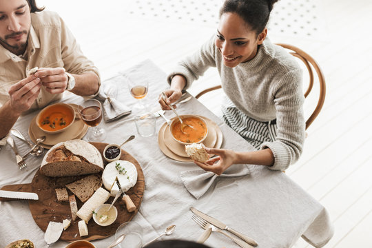 Top view of smiling african american woman sitting at the table eating cream soup with slice of bread in hand. Group of young international friends having lunch dreamily spending time in cozy cafe