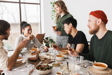 Pretty woman with wavy hair standing near table happily with colleagues. Group of young  international friends dreamily holding glasses of wine in hands having lunch together in cozy cafe