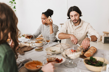 Thoughtful man with dark hair and beard sitting at the table cutting bread while afro american woman near dreamily eating. Group of young international friends together on lunch in cozy cafe