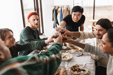 Young joyful asian man in eyeglasses and black T-shirt happily cheers glasses of wine with colleagues. Group of attractive international friends spending time together on lunch in cafe