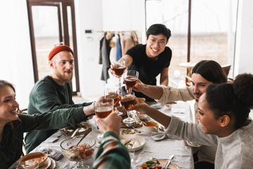 Young smiling asian man in eyeglasses and black T-shirt cheers glasses of wine with colleagues. Group of attractive international friends spending time together on lunch in cozy cafe