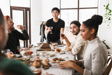 Young positive asian man in eyeglasses and black T-shirt holding glass of wine in hand happily looking aside. Group of attractive international friends spending time together on lunch in cafe
