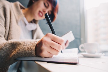 Technologies. Education. Young woman with short colorful haircut is using a smartphone and writing in a notebook while studying in the cafe