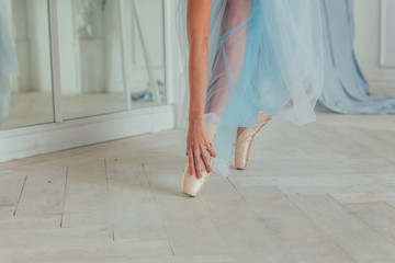 Hands of ballerina in blue tutu skirt puts on pointe shoes on leg in white light hall. Young classical ballet dancer woman in dance class