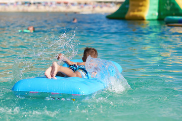 European boy is swimming in the sea on the blue air mattress, making water splashes. He is happy...