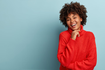 Carefree young woman with dark skin, keeps hand under chin, laughs happily, shows white teeth, wears oversized red sweater, isolated over blue background with empty space for your information