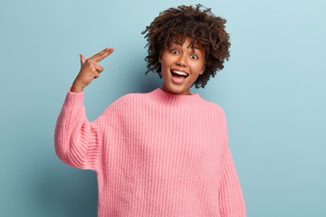 Positive ethnic woman with curly hair, dressed in pink jumper, makes suicide gesture, smiles and has fun, models over light blue background being crazy, pretends killing herself and shooting in temple