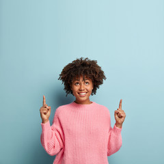 Vertical shot of good looking smiling lady with Afro haircut, wears loose pink sweater, shows...