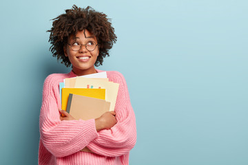 Photo of cheerful pleased schoolgirl looks upwards, dreams about recieving degree, carries papers and notepad, dressed in oversized pink jumper, isolated over blue background with copy space