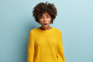 Bewildered young woman keeps jaw from astonishment, opens eyes widely, has dark skin and curly hair, wears oversized yellow jumper, isolated over blue background, cant believe in shocking revelation