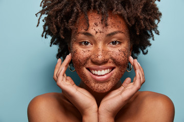 Photo of smiling black woman has cleansing mask or scrub on face, smiles broadly, enjoys beauty...