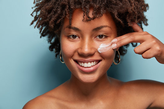 Close up shot of positive dark skinned woman with curly hair, appiles face cream on cheek, poses nude against blue background, cares of beauty, uses cosmetic product. Natural beauty concept.