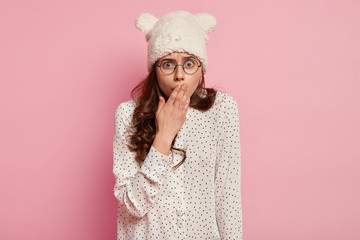 Shocked overwhelmed woman wears round spectacles, covers mouth with hand, being in stupor from shocking news, isolated over pink background. Amazed girl expresses surprisement and disbelief.