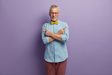 Indoor shot of handsome hesitant grandfather crosses arms over chest, points with fore fingers at...
