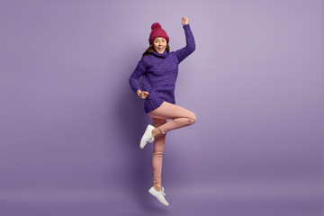 Bright image of optimistic woman feels energetic and elated, raises hands and legs, has fun indoor, wears warm clothes, isolated over purple background, rejoices good life. Optimism concept.