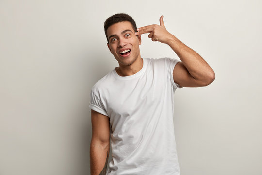 Amazed attractive young guy shoots in temple, demonstrates suicide gesture, dressed in casual t shirt, shows shooting sign, poses over white background, tired of everything, pretends he has gun