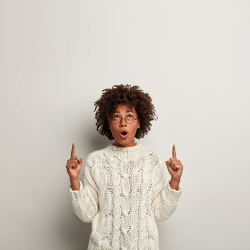 Impressed astonished dark skinned woman with crisp hair, wonder something incredible on ceiling, shows direction upwards, wears knitted sweater, isolated over white backgrund with empty space