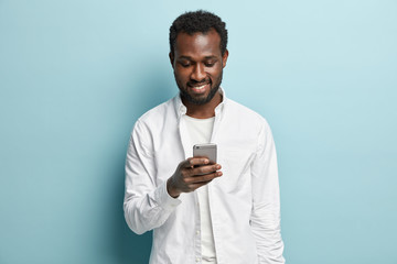 Positive black man with toothy smile, looks at smartphone, reads text message from friend, rejoices good news, wears white shirt, models in studio against blue background. Spare time and technology