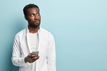 Pensive black man focused upwards, thinks what to write, has to give feedback, holds mobile phone, wears white shirt, isolated over blue studio wall with copy space, connected to wireless internet