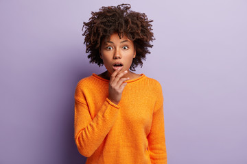 Photo of shocked lady has dark skin, keeps hand on chin, has eyes bugged, wears orange jumper, isolated over purple background, has bated breath from excitement. People and surprisement concept