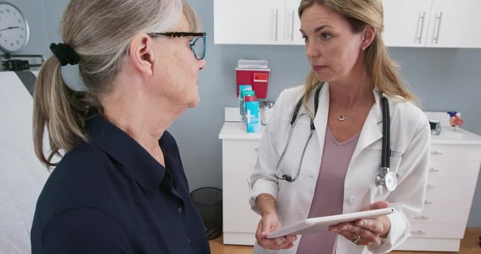 Female primary care doctor having conversation with older Caucasian woman during regular check up holding tablet computer. Senior patient talking with reassuring physician in exam room. Slow mo 4k