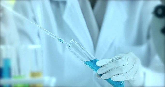 Blurred scientist doing experiment in laboratory, she dropping blue liquid into test tube, colorful test tubes on table behind him.