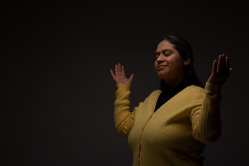 Hispanic Christian Woman Praying with Open Arms on Dark Grey Background 